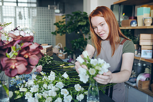 Content young florist in apron standing at counter with heap of flowers and creating floral decoration in vase