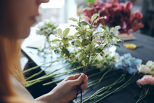 Close-up of florist standing at counter with flowers in row and adding greenery into bouquet for texture