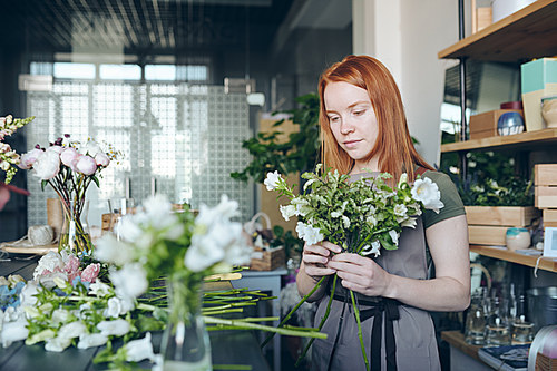 Concentrated young flower storekeeper in apron standing at counter with flowers and making tender bouquet with greenery in own shop