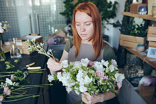 Serious creative redhead florist in apron standing at desk with tools and flowers and arranging wedding bouquet in own workshop