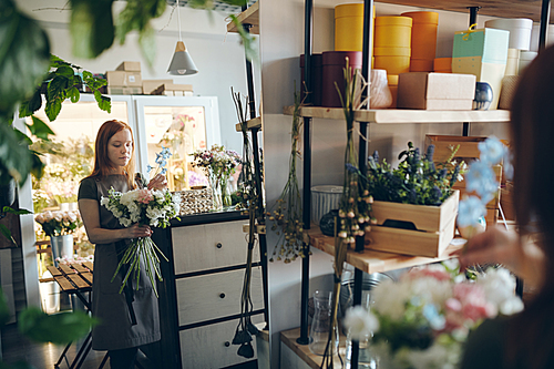 Serious busy redhead girl in apron standing in front of big mirror and finishing to arrange bouquet in flower shop