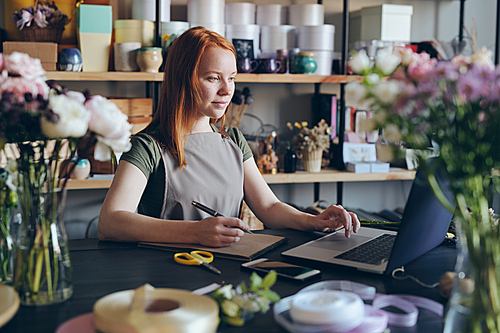 Content confident attractive young woman in apron standing at counter and using laptop while processing online orders in flower shop