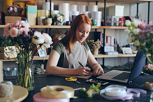 Busy young woman with red hair standing at counter and making notes in sketchpad while using laptop to buy flowers online for own store