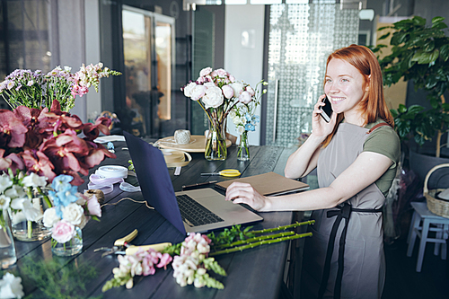 Positive young manager of flower shop standing at counter full of various flowers and using laptop while talking order by phone