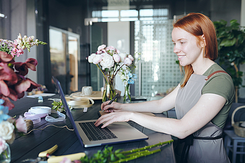 Smiling attractive female florist with red hair standing at counter with flowers and packaging items and using laptop while making list of necessary flowers