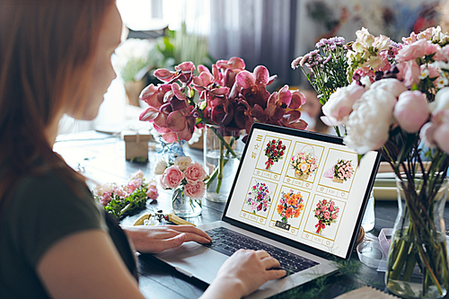 Over shoulder view of busy flower shop owner standing at counter with flowers in vases and using laptop while choosing flowers on internet