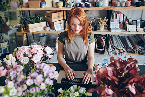 Content pretty redhead florist in apron standing at counter with flowers and working with social media of flower shop