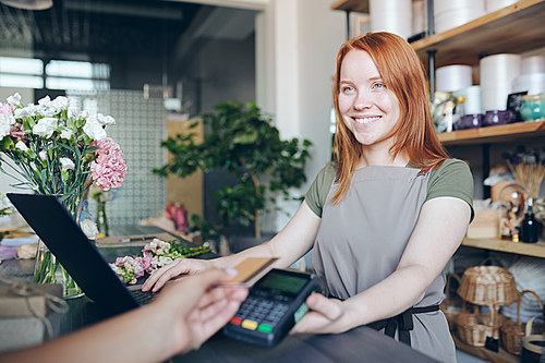 Smiling friendly redhead girl in aprin standing at counter and holding payment terminal for wireless payment while selling flowers in shop
