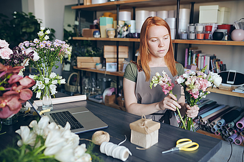 Content concentrated young redhead woman in apron standing between counter and shelves and creating beautiful floral arrangement in flower workshop
