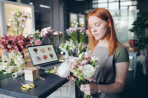 Busy attractive girl in apron standing by counter full of flowers and decoration tools and mixing flowers in bouquet while working in flower store