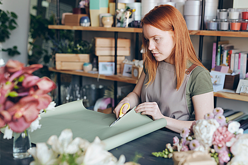 Serious busy young redhead woman in apron standing by counter and cutting wrap paper for flower packaging in flower store