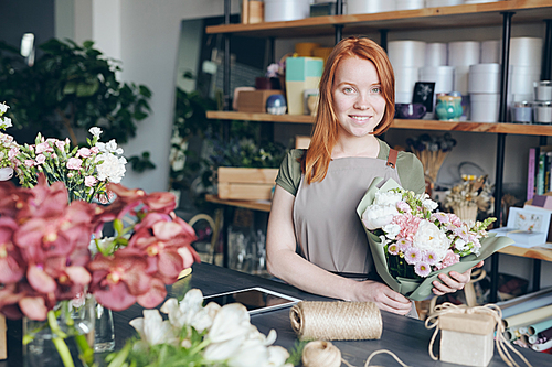 Happy attractive redhead young florist in apron holding beautiful bouquet and  while working in own shop