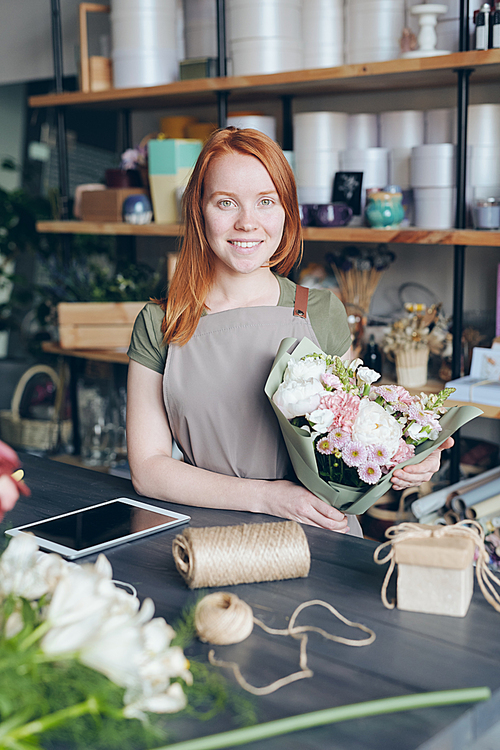 Portrait of smiling beautiful female florist with red hair standing at wooden counter with tablet, twine and flowers and holding bouquet