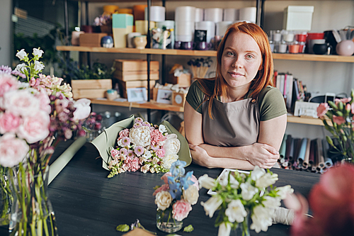 Portrait of content redhead florist in apron standing at counter with various flowers in modern flower store