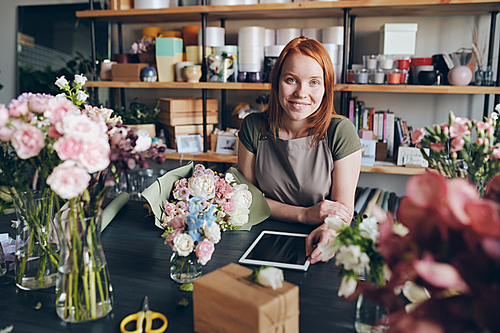Portrait of smiling beautiful young woman in apron working in cozy flower shop, she leaning on counter with flower compositions and using digital tablet