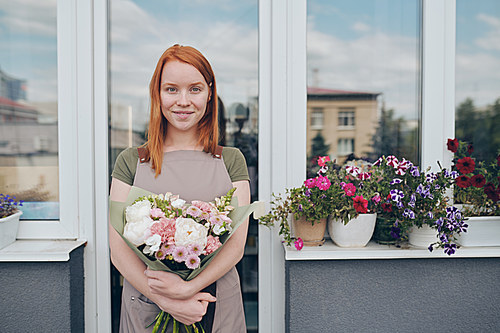 Portrait of attractive girl with red hair standing at balcony with potted flowers on window sill and embracing nice bouquet