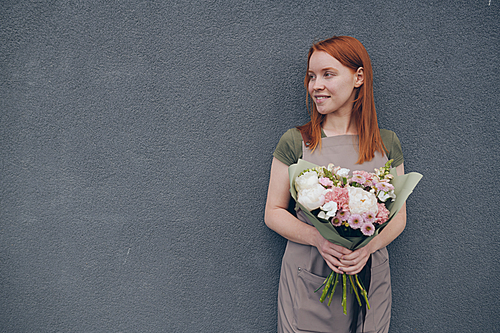 Smiling talented young female florist with red hair wearing apron standing against gray wall and holding beautiful bouquet in wrapping paper