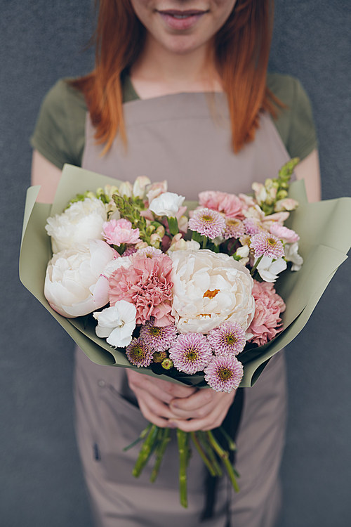 Close-up of creative florist in apron holding beautiful bouquet with peonies wrapped into green paper