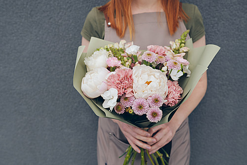 Close-up of unrecognizable florist in apron standing against gray wall and holding bright tender bouquet in green kraft paper