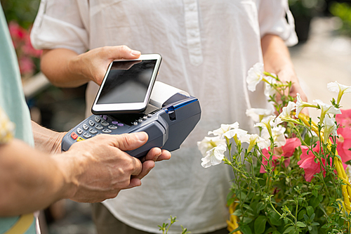 Young client of flower shop or garden center holding smartphone over payment machine while buying flowers