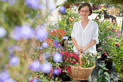 Pretty brunette female gardener with basket of flowers looking at you while standing inside garden center