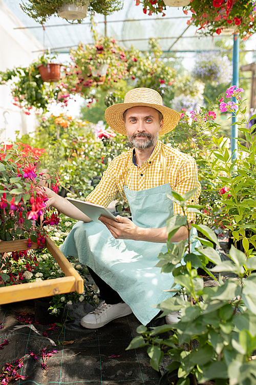 Contemporary farmer in workwear looking at you while searching for names of new sorts of flowers in greenhouse