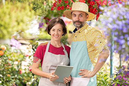Two successful farmers in workwear  while standing among blooming flowers in greenhouse