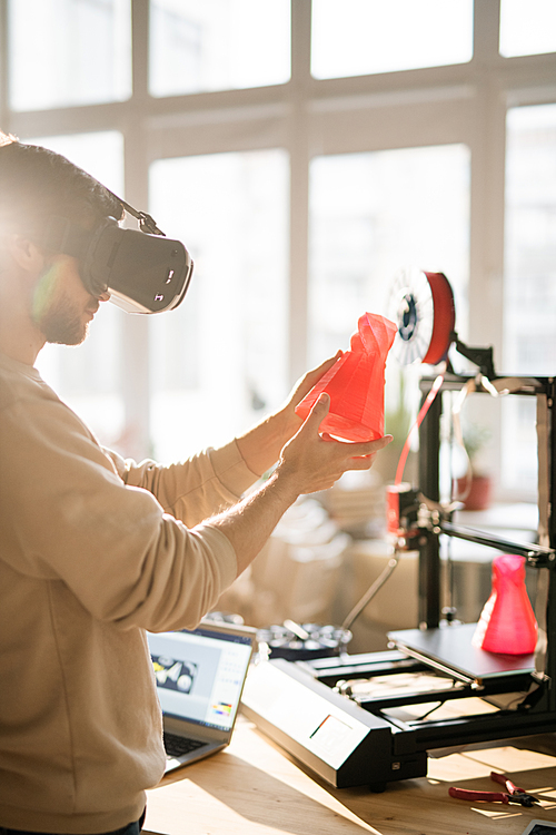Young businessman in vr headset holding red plastic object in front of himself while going to put it on 3d printer in office