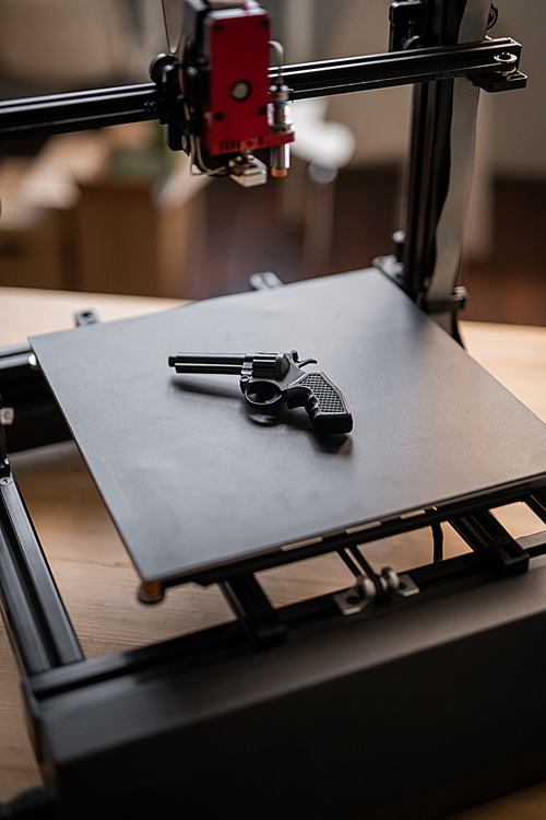 Black revolver gun lying on working surface of modern 3d printer with printhead hanging above