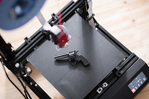 Overview of black revolver gun on working surface of 3d printer on wooden table inside modern office