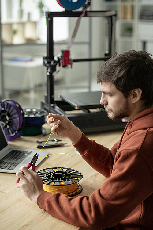 Creative man sitting by table and cutting piece of yellow filament while going to make 3d printing of new object