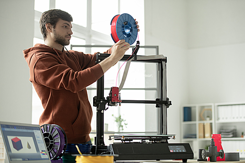 Young man putting new spool with red filament in 3d printer while going to print geometric figures