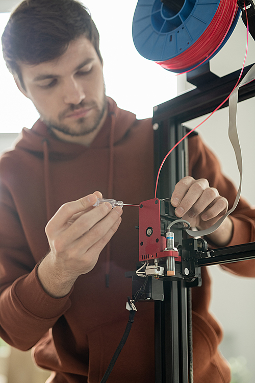 Young technician with screwdriver repairing printhead or fixing one of panels of 3d printer in office