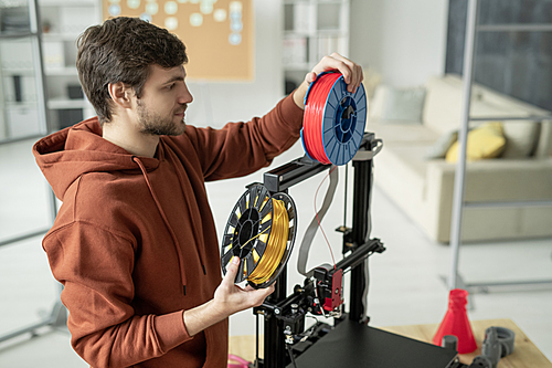 Young creative man changing spool with filament while standing by 3d printer before printing objects of various colors