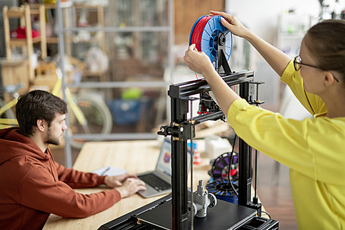 Young female office worker changing spool with red filament on 3d printer with colleague networking on background
