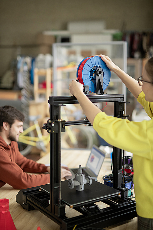 Young woman putting new spool with red filament on 3d printer while her colleague searching for online ideas on background