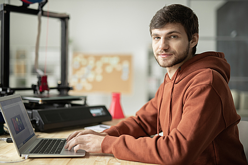 Bearded guy in brown hoodie sitting in front of laptop in office while working over sketches of items for 3d prirnting