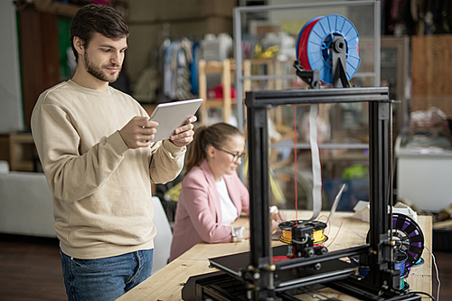 Young man with touchpad looking at online figure on display while standing in front of 3d printer on background of colleague