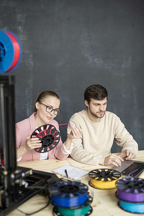 Young female looking at red filament on spool while sitting next to her colleague surfing for websites in front of laptop