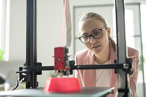 Young businesswoman standing in front of 3d printer in process of creating pink object in office
