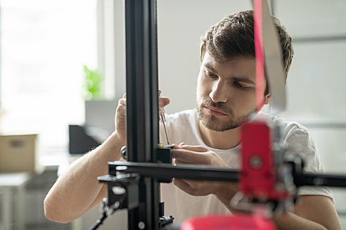 Young businessman standing in front of 3d printer while fixing some details before start of work