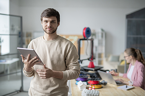 Young businessman with touchpad standing in front of camera on background of colleague working with 3d printer