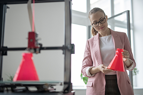 Elegant female designer holding just printed red object of geometric shape while standing by 3d printer