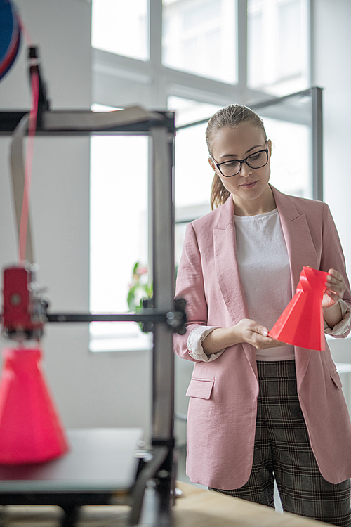 Elegant young designer with just printed red object of geometric shape standing by workplace in office