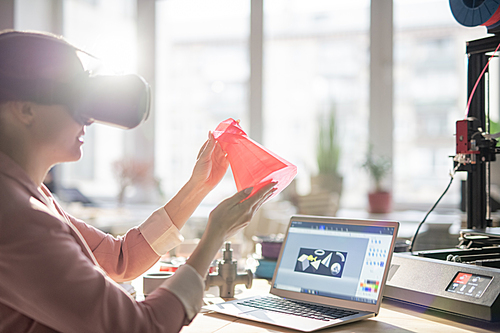 Contemporary young businesswoman in vr headset holding printed 3d object in front of herself while sitting by workplace