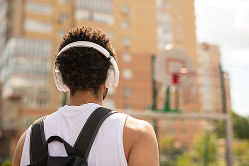 Rear view of young intercultural sportsman with headphones and backpack standing on playground in urban environment