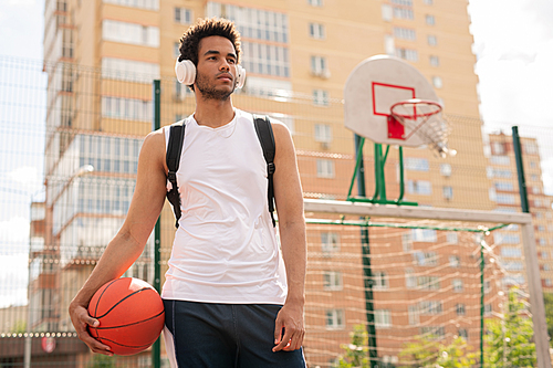Sportive guy with ball for playing basketball listening to music in headphones on playground of field