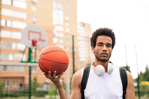 Young serious active male basketballer in white t-shirt holding ball while standing in front of camera on playground