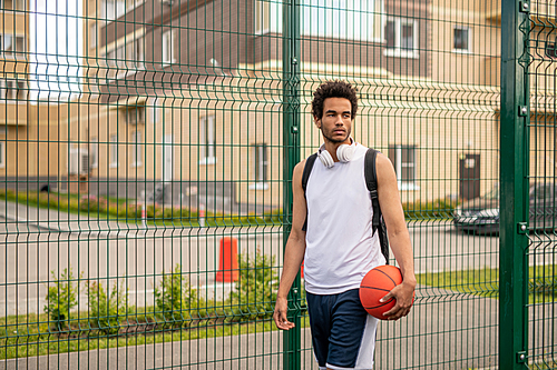 Young mixed-race professional basketball player with ball standing by fence of playground in urban environment