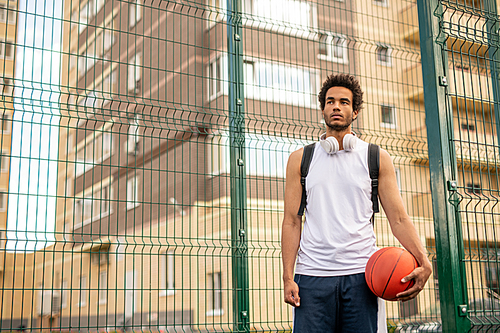 Young sportsman in white t-shirt with ball for playing basketball standing by fence surrounding court